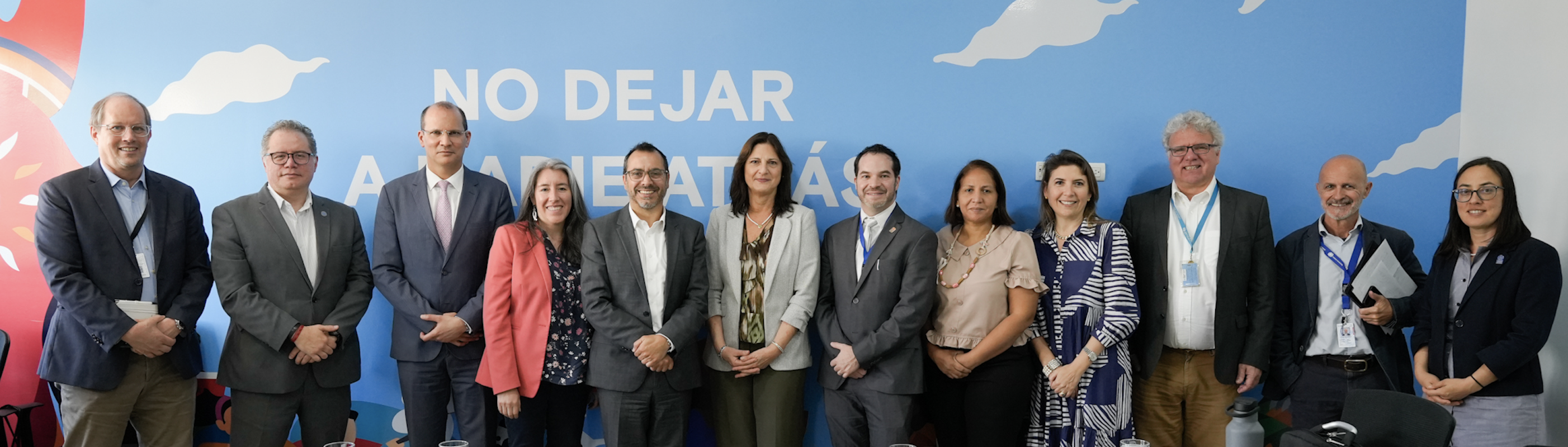 La imagen muestra a las y los representantes del Equipo País de las Naciones Unidas en Honduras (UNCT, por sus siglas en inglés) posando juntas frente a una pared azul con el lema “NO DEJAR A NADIE ATRÁS”, en letras grandes y blancas. Las y los integrantes están vestidos formalmente y se ubican en una sola fila, sonriendo a la cámara.
