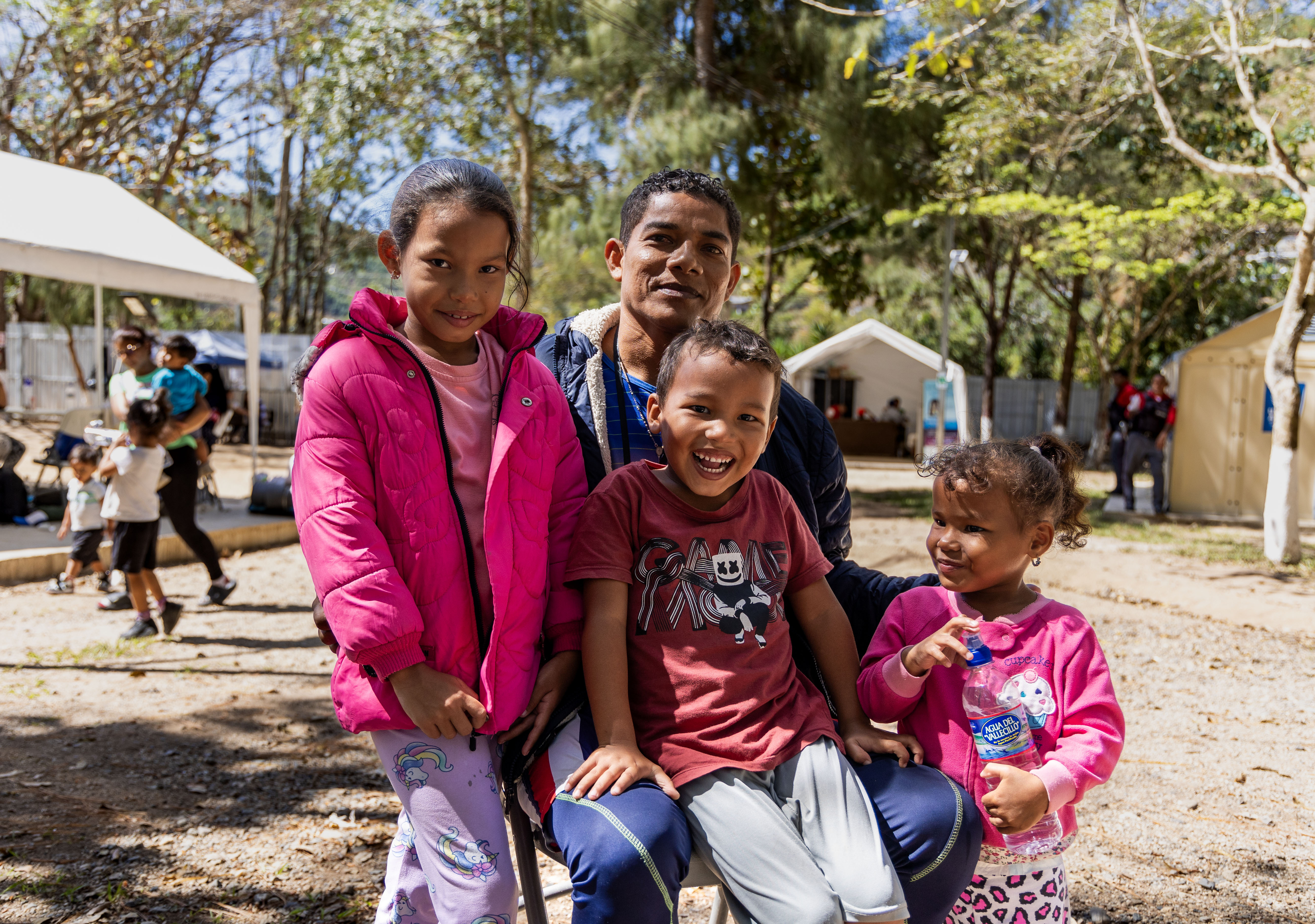 La imagen muestra a un hombre adulto sentado, rodeado de cuatro niños, dos niñas y dos niños, en un ambiente al aire libre y soleado. Todos están sonriendo o con expresiones alegres, reflejando un momento de cercanía y calidez familiar. Al fondo se observan carpas blancas, árboles y otras personas caminando o interactuando, lo que sugiere que se trata de un espacio comunitario o un albergue temporal.