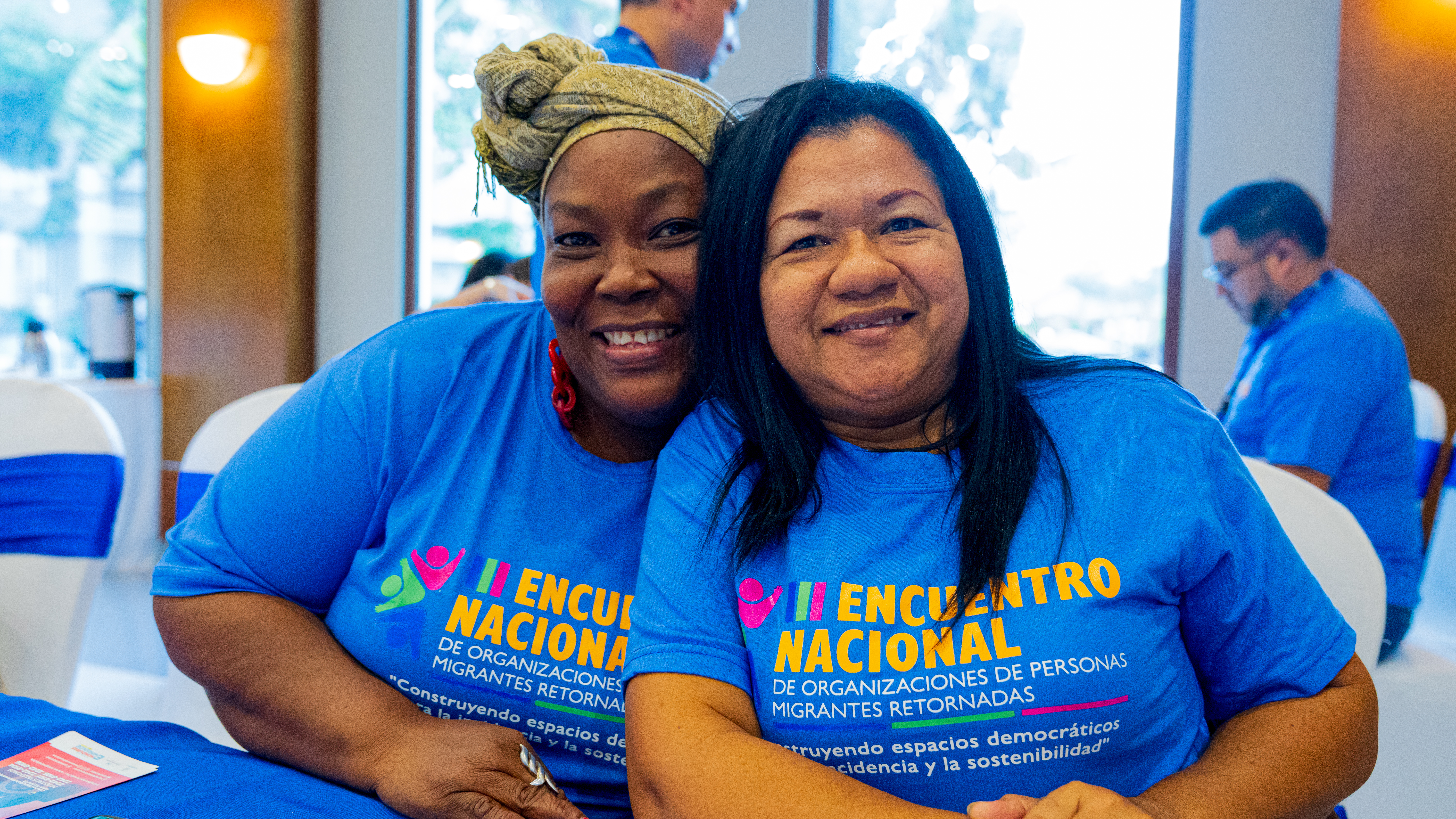 La imagen muestra dos mujeres adultas sentadas y abrazadas, sonriendo a la cámara. Ambas visten camisetas azules del "II Encuentro Nacional de Organizaciones de Personas Migrantes Retornadas". El ambiente es alegre dentro de un espacio cerrado y bien iluminado, con más personas participando en el fondo. La escena transmite unidad, empoderamiento y trabajo colectivo por los derechos de las personas migrantes.