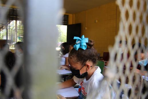 Se observa a través de una ventana a una niña con mascarilla escribiendo en su cuaderno dentro de un aula de clase