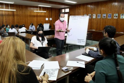 Joven hombre modera taller. Se le ve hablando con una de las personas que se sienta en la mesa tipo U que está en el salón. A su lado una joven mujer toma notas en una computadora.