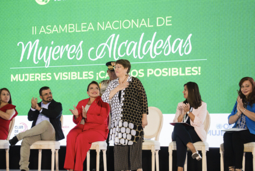 En la imagen, siete personas participan en un evento oficial sobre un escenario. En el centro se encuentra la primer mujer presidenta de Chile, Michelle Bachelet, de pie con la mano en el pecho, mientras las demás personas aplauden desde sus asientos a su alrededor. El fondo verde muestra el texto: “II Asamblea Nacional de Mujeres Alcaldesas,  Mujeres visibles ¡cambios posibles!”. El ambiente es solemne y de reconocimiento.