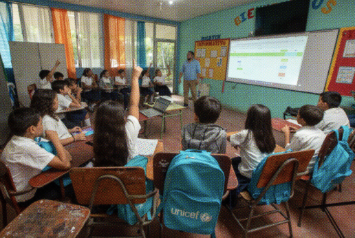 La imagen muestra un aula escolar donde un grupo de niñas y niños están sentados en círculo prestando atención a un maestro que se encuentra de pie frente a una pizarra digital proyectada. Algunos estudiantes tienen la mano levantada para participar. Todos visten uniforme escolar blanco y portan mochilas celestes con el logotipo de UNICEF, colocadas en el respaldo de sus sillas. El aula está decorada con cortinas de colores y carteles escolares, creando un ambiente dinámico y participativo.
