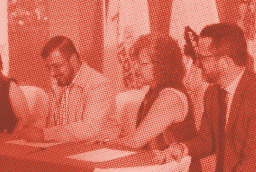 Imagen en tonos rojos que muestra a dos mujeres y dos hombres sentados en una mesa, participando en un acto formal. Al fondo se observan varias banderas, entre ellas la de la Asociación de Municipios de Honduras (AMHON), Naciones Unidas y ONU Mujeres.