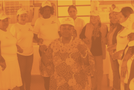 Imagen en tonos anaranjados que muestra a un grupo de mujeres reunidas en círculo en un espacio cerrado. Varias de ellas visten camisetas blancas y gorras, mientras que una mujer en el centro, con ropa estampada.