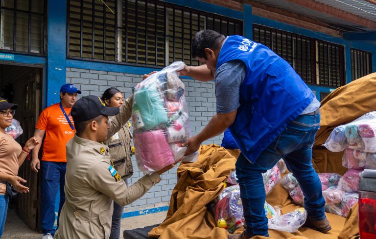 La imagen muestra a varias personas colaborando en la entrega de ayuda humanitaria. Un hombre con chaleco azul de la OIM (Organización Internacional para las Migraciones) pasa un paquete grande de víveres y productos de higiene a un miembro uniformado de una institución local. Al fondo, otras personas también participan en la actividad frente a un edificio de ladrillo gris con ventanas enrejadas.